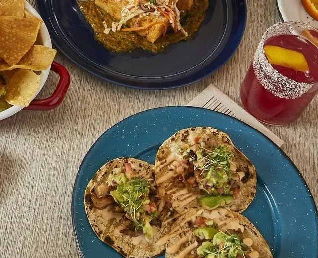 Shared plates at Barrio Costero in Asbury Park: grilled tacos topped with avocado and microgreens on a blue plate, chips, an enchilada in salsa verde, and a hibiscus cocktail