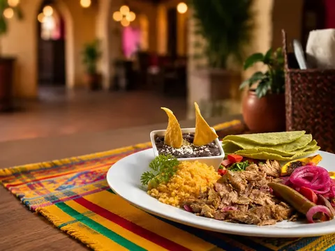 Plated Oaxacan dish of shredded meat with yellow rice, black beans, pickled onions and tortillas on a colorful woven table runner, with the warmly lit arched dining room of Fiesta Oaxaca behind