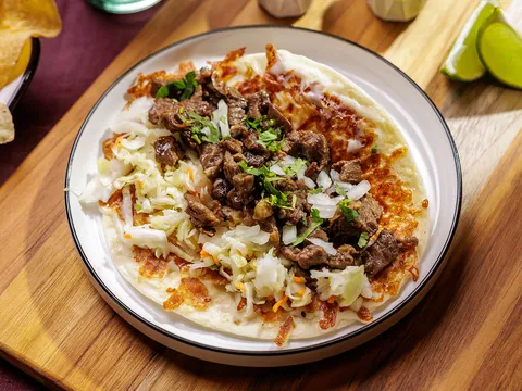 A Salsa Limón taco 'con todo' — grilled asada with crispy melted cheese, cabbage, onion and cilantro on a flour tortilla, served with lime wedges and tortilla chips on a wooden board