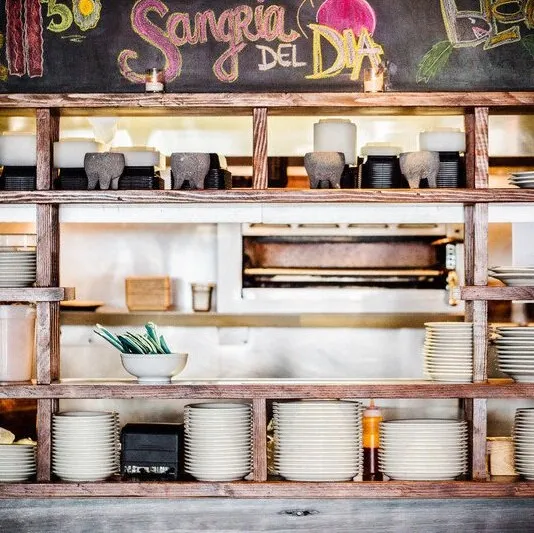 Rustic wooden shelving stacked with plates below a hand-chalked "Sangria del Día" board inside Bodega Taco Bar in Fairfield, Connecticut